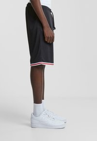 Pantalones cortos deportivos negros con cintura ajustable, con rayas rojas y blancas en el dobladillo, combinados con zapatillas blancas y calcetines.
