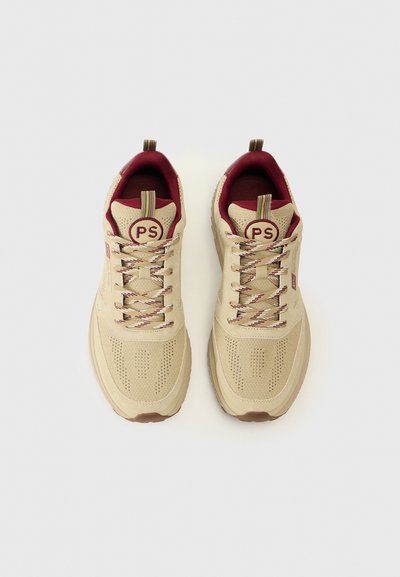 Baskets beiges avec des accents bordeaux, texture perforée et lacets beiges. Présente un logo rond sur la languette et une languette de tirage multicolore.