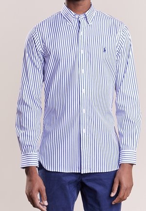 Mężczyzna ubrany w białą koszulę zapinaną na guziki z niebieskimi pionowymi paskami oraz niebieskie spodnie, stojący na jednolitym tle.