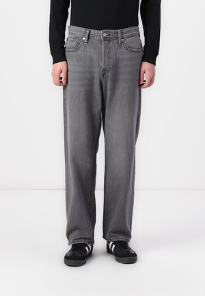 Osoba ubrana w luźne, wyblakłe szare dżinsy, czarną bluzkę z długim rękawem oraz czarne tenisówki z białymi paskami, stojąca na białym tle.