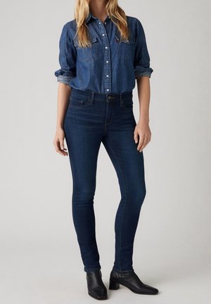 Femme portant une chemise en jean bleu foncé boutonnée avec les manches retroussées, un jean skinny bleu foncé et des bottines noires à talons, debout devant un fond gris.