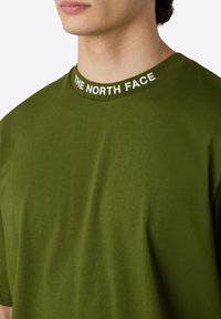 Olivengrünes T-Shirt mit kurzen Ärmeln und rundem Halsausschnitt, das mit einem weißen Logo-Band versehen ist. Weiche Textur, normale Passform und schlichtes Design.