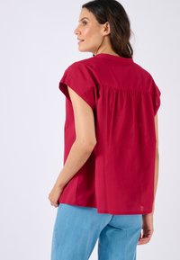 Top rosso a maniche corte con vestibilità loose e dettaglio arricciato sul retro. Tessuto liscio di un colore uniforme, abbinato a jeans blu.