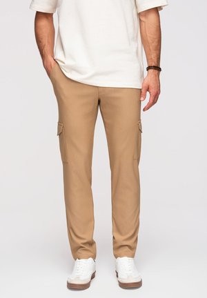 Mężczyzna w beżowych spodniach typu cargo, białych trampkach i białej koszuli z krótkim rękawem, z jedną ręką w kieszeni.