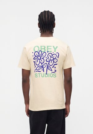 Muž s vrkočmi, ktorý nosí béžové tričko so zeleným nápisom "OBEY STUDIOS" a modrým abstraktným kvetinovým motívom na chrbte, čierne nohavice.
