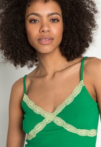 Camisola sem mangas verde com nervuras, decorada com uma bainha de rendinha verde claro que forma um X no peito. Possui alças finas ajustáveis.