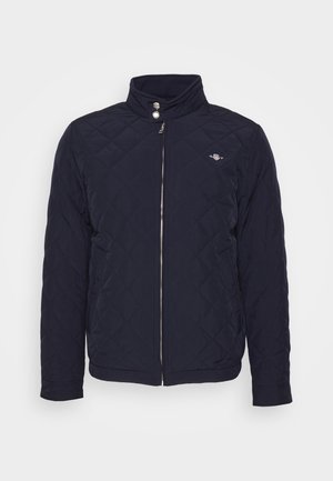 Navy quilted jacket with a high collar, zip closure, and press-stud detailing; features a small logo on the chest. Soft, smooth texture.