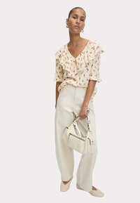 Bloemige blouse met ruches in crème, hooggeplaatste witte broek en een crèmekleurige handtas met accenten. Platte beige schoenen maken de outfit compleet.