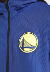 Veste bleue zippée avec le logo des Golden State Warriors représentant un pont stylisé et un ballon de basket au niveau de la poitrine.