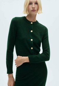 Vihreä neulecardigan, jossa on kultaiset napit, pyöreä kaula-aukko, pitkät hihat ja pehmeä texture. Yksinkertainen, istuva malli, jota wearataan yhteensopivan mekon päällä.