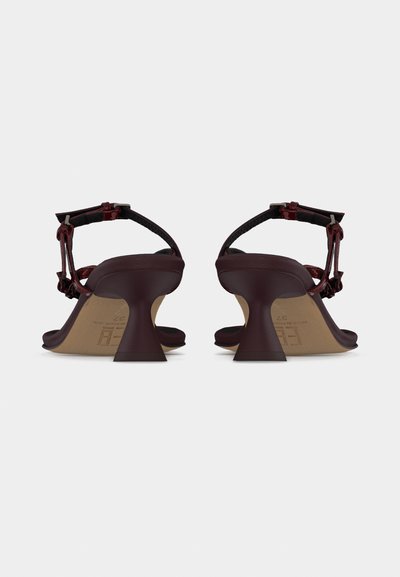 Ζευγάρι σκούρα κόκκινα πέδιλα με ψηλό τακούνι και λουράκια στον αστράγαλο, εμφανιζόμενα από πίσω σε απλό ανοιχτό χρωματικό φόντο.