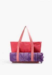 Sac cabas rouge avec poignées roses maintenant une serviette violette roulée ornée d'un motif floral, posé sur un fond blanc.
