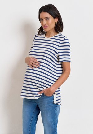 Zwangere vrouw met donker haar die een wit en marineblauw gestreept T-shirt met korte mouwen en een blauwe spijkerbroek draagt, haar buik vasthoudend en licht glimlachend.