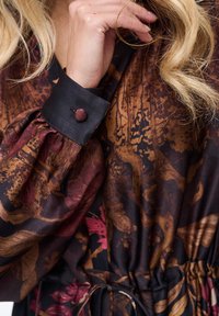 Sukienka z długim rękawem z ciemnym kwiatowym nadrukiem w kolorach rdzawego, złotego i czarnego. Posiada czarny mankiet z okrągłym ozdobnym guzikiem oraz marszczoną talię.