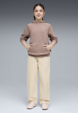 Jeune femme debout, mains dans la poche d'un sweat à capuche beige, portant un pantalon large crème et des baskets rose clair sur fond gris.