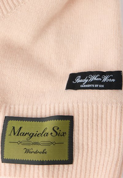 Világos rózsaszín kötött ruhadarab bordázott textúrával; fekete és zöld címkékkel, melyeken a „Margiela Six” és a „Ready When Worn” felirat olvasható.