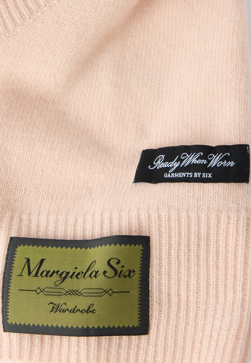 Lyserød strikket beklædningsgenstand med ribbet struktur; har sorte og grønne mærker med teksten "Margiela Six" og "Ready When Worn."