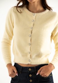 Suéter de cardián amarillo suave con cuello redondo, cierre de botones con acentos en tono plateado y textura suave. Se combina con denim oscuro.