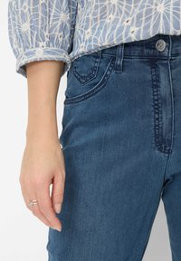 Dark blue denim jeans featuring stitched pockets and a button closure. A hand with a silver ring rests against the fabric, highlighting texture.