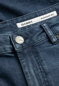 Σκούρο μπλε τζιν με ασημί κουμπί και ετικέτα GABBA με κεντημένο το μήνυμα "ΣΥΝΑΦΕΣ ΑΠΟ ΤΟ 1983" κάτω από αυτήν. Κοντινή λήψη της μέσης.