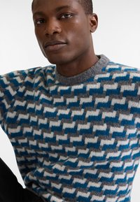 Maglione lavorato a maglia con un motivo geometrico nei colori blu, grigio e bianco. Presenta un collo grigio arrotondato e un tessuto testurizzato con cuciture visibili.