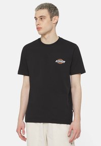 Camiseta de algodón negra con un pequeño logo naranja y blanco de "Dickies" en el lado izquierdo del pecho. Diseño de manga corta y cuello redondo.