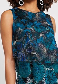 Top sin mangas de tela translúcida con un patrón floral y de mariposas en tonos azules. El diseño incluye capas y un escote redondo.