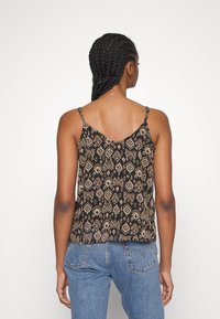 Woman with braided hair wearing a black patterned spaghetti strap top and blue jeans, facing away against a plain white background.