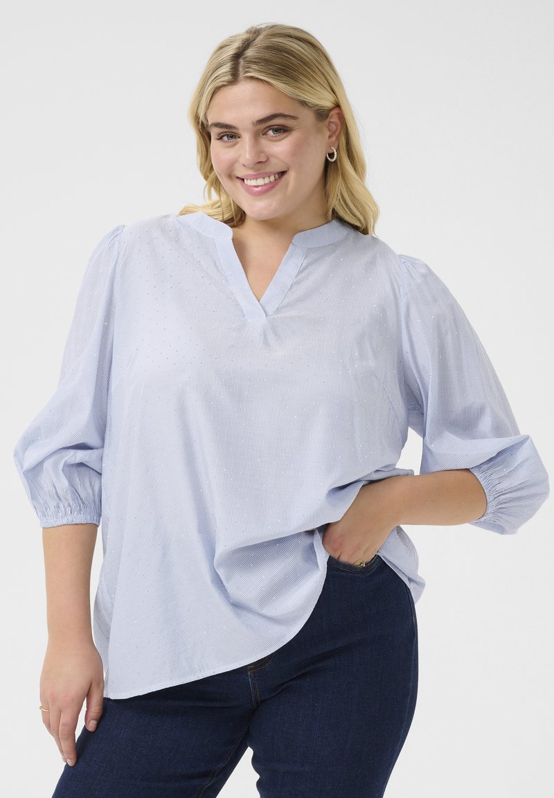Blusa azzurro chiaro a maniche lunghe con scollo a V e maniche a sbuffo. Presenta un motivo testurizzato e strisce sottili. Abbinata a jeans scuri.