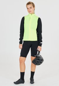 Neon gele fietsjack met zwarte lange mouwen, gecombineerd met zwarte shorts en zwarte fietsen schoenen. Houdt een zwarte helm vast.