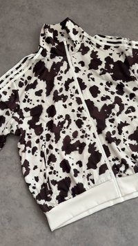 White and black cow print track jacket with a front zipper, zippered side pocket, and three white stripes on the shoulders and sleeves.