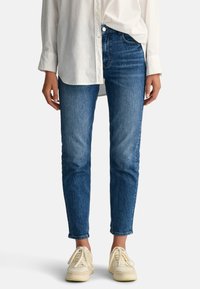 Fitte blauwe denim jeans met een lichte vervaging, een middelhoge taille en een vijf-pocket ontwerp, gecombineerd met witte sneakers en een los wit shirt.