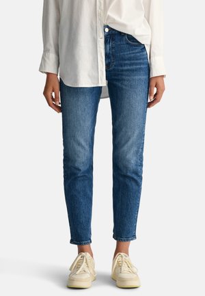 Fitted blue denim jeans with a slight fade, a mid-rise waist, and five-pocket design, paired with white sneakers and a loose white shirt.