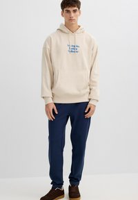 Joven de pie con sudadera beige con texto azul "Los Angeles Heritage Collective", pantalones azul marino y zapatillas marrones sobre fondo blanco.
