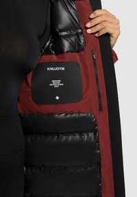 Veste matelassée avec une doublure intérieure noire et un tissu extérieur rouge profond. Étiquette de la marque visible dans la poche intérieure, avec un détail zippé.