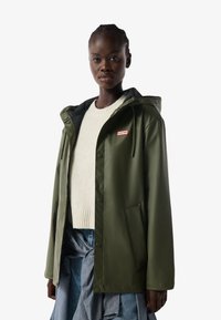 Mujer negra con una chaqueta impermeable verde de Hunter sobre un suéter blanco y una falda azul, de pie frente a un fondo blanco.