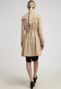 Abrigo beige con cintura ajustada, espalda plisada y mangas largas. Detallado con un amplio cuello y botones negros.