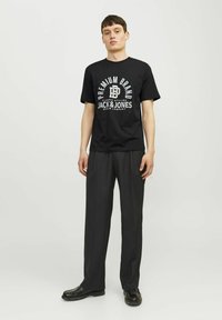 Juodas marškinėliai su balta grafika, "PREMIUM BRAND JACK & JONES." Suderinti su tamsiomis kelnėmis ir juodais batais, nustatyti neutralioje aplinkoje.
