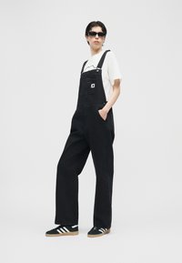 Άτομο που φοράει μαύρη τζιν σαλοπέτα, λευκό μπλουζάκι, μαύρα γυαλιά ηλίου και μαύρα αθλητικά παπούτσια Adidas με λευκές ρίγες, στέκεται μπροστά σε απλό φόντο.