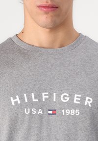 Sivi pamučni majica s kratkim rukavima i okruglim izrezom, s natpisom "HILFIGER USA 1985" otisnutim u bijeloj boji i malim logotipom, glatke teksture.