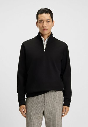 Schwarzer Reißverschluss-Hoodie mit hohem Kragen, aus glattem Stoff. Getragen über einem Hemd mit Kragen und kombiniert mit karierten Hosen.