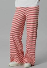 Persona che indossa sneakers bianche e pantaloni larghi a righe orizzontali rosse e bianche, in piedi contro uno sfondo grigio semplice.