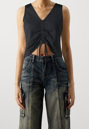 Frau trägt ein schwarzes, gerafftes, ärmelloses Crop-Top und hoch taillierte, ausgewaschene Cargo-Jeans mit mehreren Taschen.