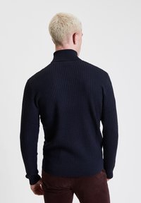 Maglione lavorato a maglia di colore blu scuro con colletto alto, superficie testurizzata e polsini a costine. Presenta un design aderente e una finitura liscia e morbida.