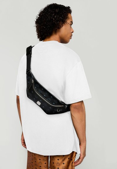 Borsa a tracolla nera con motivo testurizzato, tracolla regolabile e chiusura a zip. Indossata sopra una camicia bianca con pantaloni corti a motivo marrone.