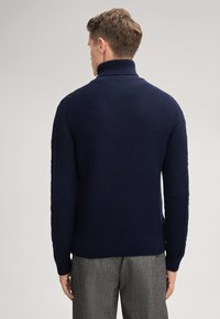 Mann mit kurzen braunen Haaren, der einen navyblauen gerippten Rollkragenpullover und graue Hose trägt, steht mit dem Rücken zu einem schlichten Hintergrund.