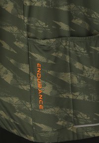 Tissu à motif camouflage vert avec une texture en mesh, une poche cousue et un texte vertical orange "ENDURANCE".