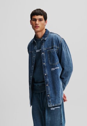 Denim jakna u srednje plavoj boji s opuštenim krojem, otvaranjem na gumbe, jednim džepom na prsima i diskretnim brendiranjem. U kombinaciji s odgovarajućim plavim duksericom.