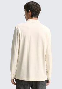 Cremefarbener Langarm-Polohemd mit strukturierter Oberfläche, ausgestattet mit einem klassischen Kragen und geradem Saum. Schlichtes Design mit minimalen Nähdetails.