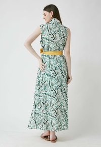 Maksikleita ar zaļu un baltu abstraktu rakstu, bez piedurknēm, un dzeltenu jostu viduklī. Maigi plūst līdz grīdai.